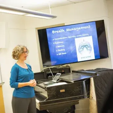 a professor lectures about vocal health with an anatomy chart behind her