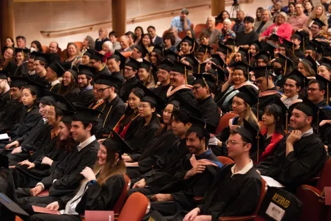 students sit in the theater wearing their regalia